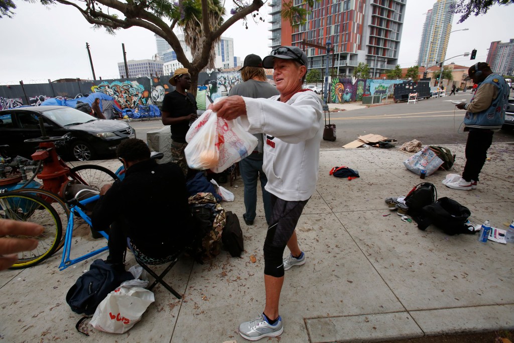 Triangle Project volunteer Bobbi Sherg, 60, cheerfully greets people camping out on the sidewalk near the intersection of 16th Street and National Avenue on Monday, June 13, 2022. Sherg retired from FedEx several years agon and has been volunteering with projects that assist the homeless in San Diego. / Photo by Peggy Peattie for Voice of San Diego