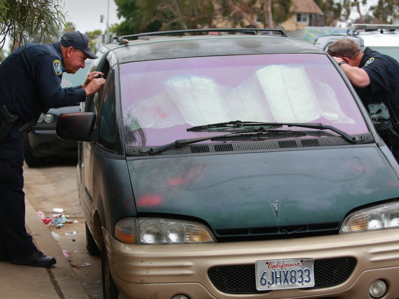 San Diego police officers peer into a partially open window of a van parked along 17th Street near Logan Avenue just after sunrise, Monday, June 13, 2022. Officers would eventually call a tow truck and have the vehicle towed, even though the owners were sleeping in a tent less than 20 feet away. / Photo by Peggy Peattie for Voice of San Diego