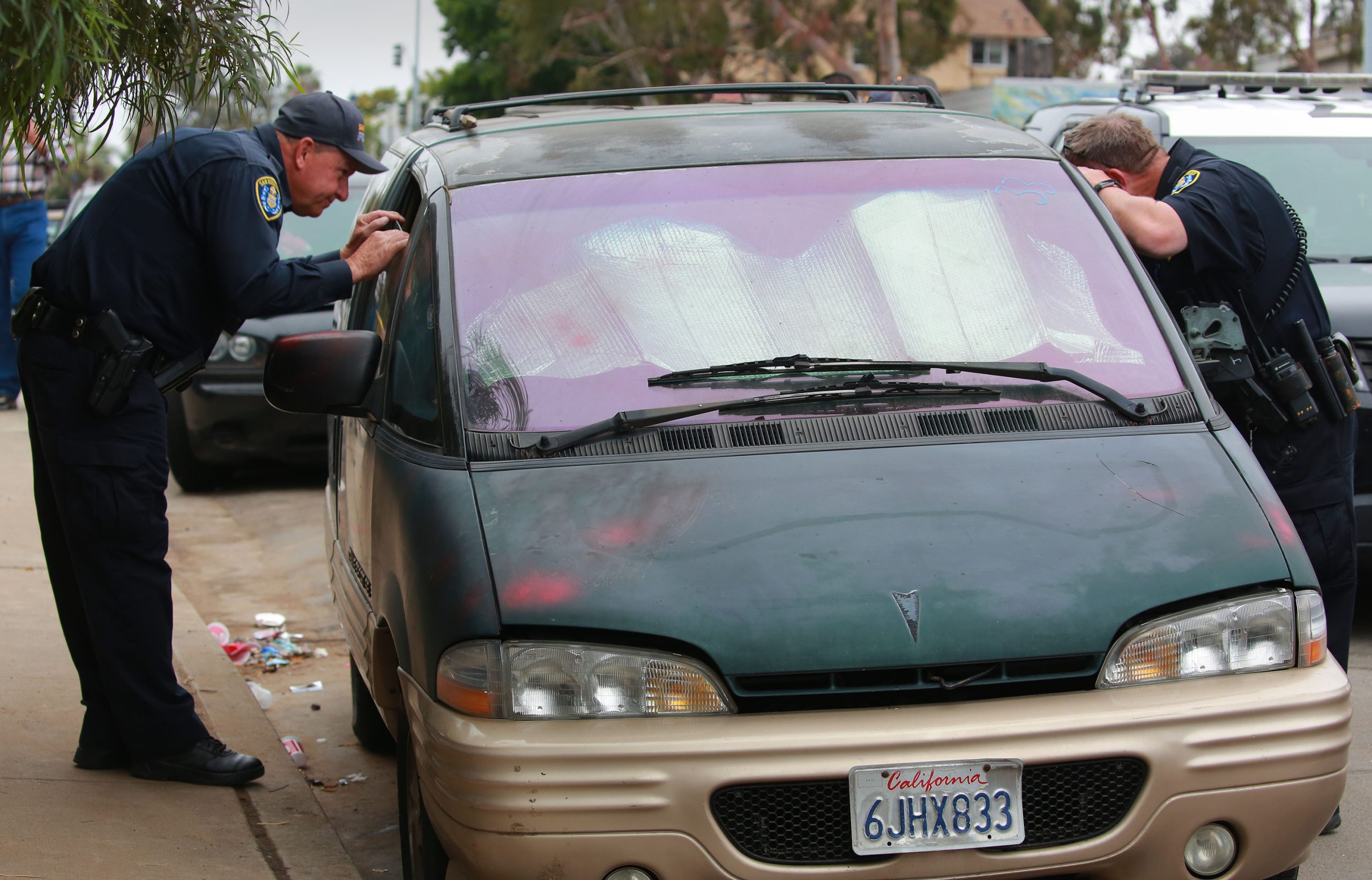 San Diego police officers peer into a partially open window of a van parked along 17th Street near Logan Avenue just after sunrise, Monday, June 13, 2022. Officers would eventually call a tow truck and have the vehicle towed, even though the owners were sleeping in a tent less than 20 feet away. / Photo by Peggy Peattie for Voice of San Diego
