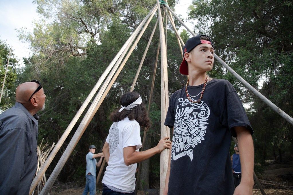 A teenager looks for the men carrying the next teepee pole to be added to the frame, while Guillermo Aranda surveys the group's progress./ Photo by Jakob McWhinney
