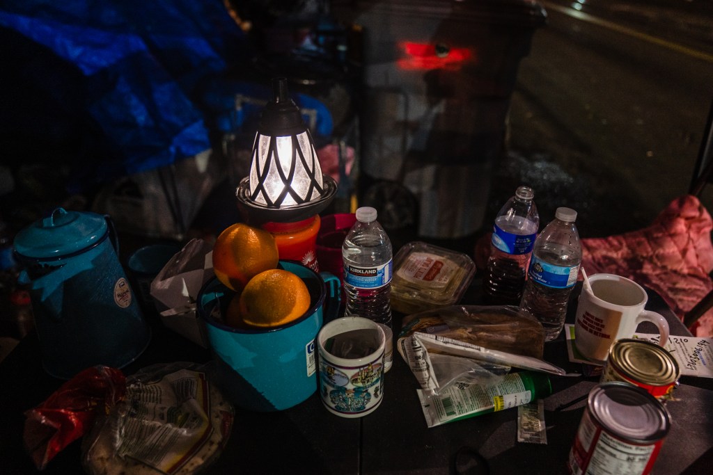 A solar light along with oranges, bottles of water, canned food sit on a table at a homeless encampment on Commercial Street in downtown on March 30, 2023.