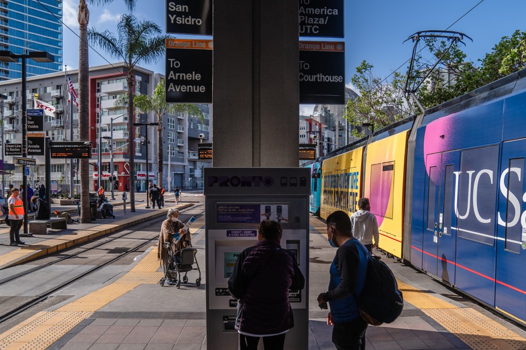 A man helps an older woman use the PRONTO machine so she can purchase a ticket to ride the trolley at 12th and Imperial Avenue in downtown on May 1, 2023.