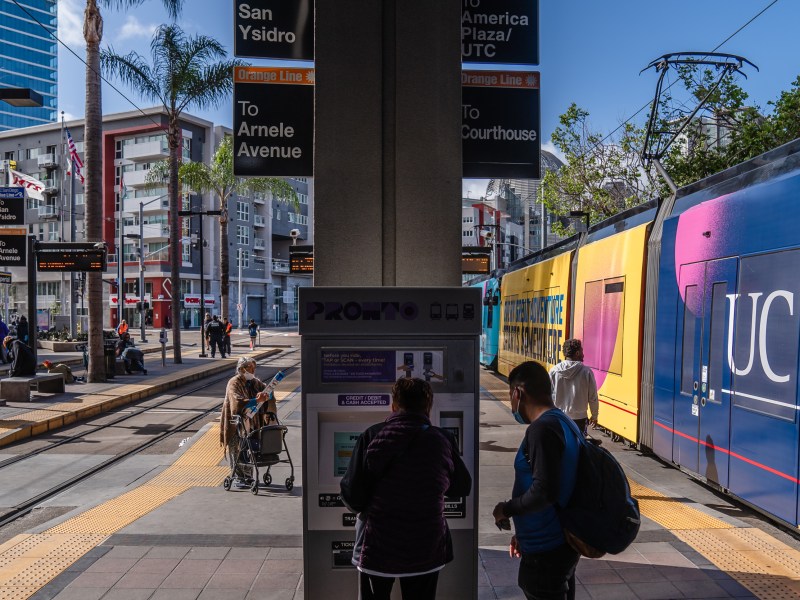 A man helps an older woman use the PRONTO machine so she can purchase a ticket to ride the trolley at 12th and Imperial Avenue in downtown on May 1, 2023.