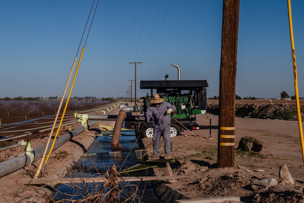 A worker cleans grates in an irrigation ditch in Imperial Valley on Oct. 10, 2023.. A pump sends Colorado River water to sprinklers on a farm field.