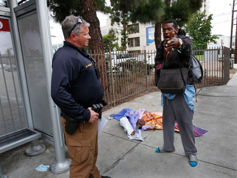 Christopher Pair, 45, right, tells San Diego police Officer Michael Padgett, left, that he spent the night behind a bus stop near UCSD Medical Center in Hillcrest after being discharged overnight when buses weren’t running. Pair said he’d take a shelter bed when Padgett offered to find one but the officer ultimately learned there weren’t any available beds. / Photo by Peggy Peattie for Voice of San Diego