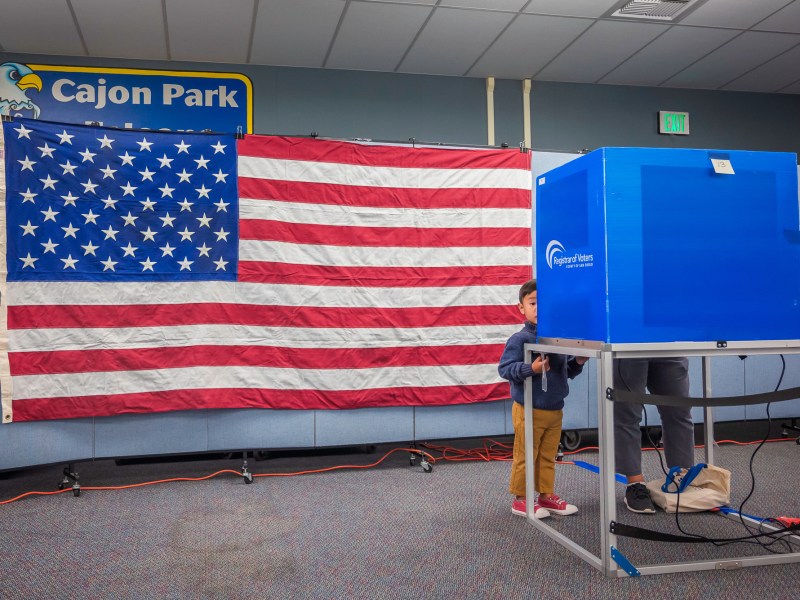 A little boy stands near a voting both, while his guardian votes on Tuesday, Nov. 5, 2024. / Photo by Vito di Stefano for Voice of San Diego