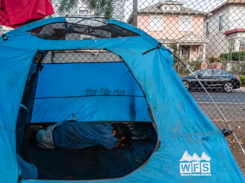 A man rests in a tent at a homeless encampment next to Interstate 5 North ramp near downtown San Diego on July 15, 2025. / Ariana Drehsler for Voice of San Diego